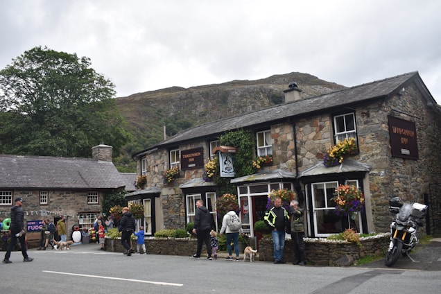 A quaint stone inn adorned with colorful flowers is set against a hilly backdrop. People are gathered around, some walking dogs while others are engaged in conversation. A motorcycle is parked nearby, adding a touch of modernity to the rustic scene. The atmosphere is lively, with a sense of community.