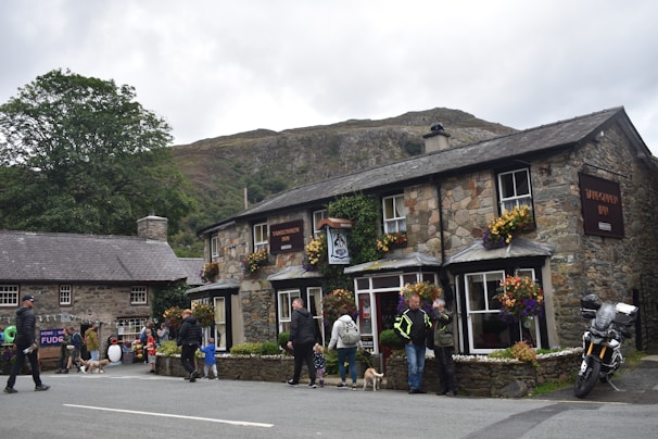 A quaint stone inn adorned with colorful flowers is set against a hilly backdrop. People are gathered around, some walking dogs while others are engaged in conversation. A motorcycle is parked nearby, adding a touch of modernity to the rustic scene. The atmosphere is lively, with a sense of community.