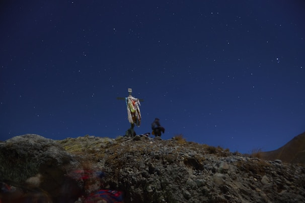 A cross adorned with colorful fabric stands on a rocky terrain under a starry night sky. A figure appears to be moving or blurred near the cross, adding a sense of mystery.