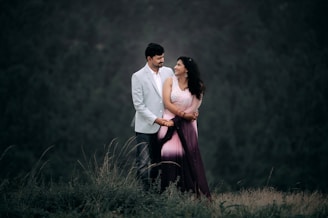 A man and a woman stand together outdoors, with the man wearing a light gray suit and the woman dressed in a pink and purple sari. They are looking at each other affectionately, surrounded by greenery with a blurred forest background, creating a romantic and serene atmosphere.