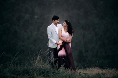 A man and a woman stand together outdoors, with the man wearing a light gray suit and the woman dressed in a pink and purple sari. They are looking at each other affectionately, surrounded by greenery with a blurred forest background, creating a romantic and serene atmosphere.