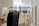 A clothing rack filled with neatly hung shirts, primarily in neutral colors like white and black, within a minimalist and well-lit interior space. The background includes soft lighting and modern architectural details such as arches and grid-patterned glass blocks.