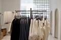 A clothing rack filled with neatly hung shirts, primarily in neutral colors like white and black, within a minimalist and well-lit interior space. The background includes soft lighting and modern architectural details such as arches and grid-patterned glass blocks.
