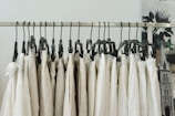 A series of beige and white clothing items hanging on black plastic hangers from a metal garment rack. The background has a blurred image of a floral poster and a part of an architectural structure.