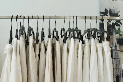 A series of beige and white clothing items hanging on black plastic hangers from a metal garment rack. The background has a blurred image of a floral poster and a part of an architectural structure.