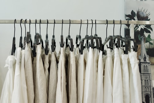 A series of beige and white clothing items hanging on black plastic hangers from a metal garment rack. The background has a blurred image of a floral poster and a part of an architectural structure.