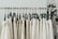 A series of beige and white clothing items hanging on black plastic hangers from a metal garment rack. The background has a blurred image of a floral poster and a part of an architectural structure.