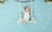 A baby dressed in a white outfit lies on a decorative swing made of white ropes and a wooden branch. The background is light blue with scattered small white flowers. Green leaves and flowers decorate the top corners, enhancing the whimsical and serene atmosphere.