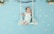 A baby dressed in a white outfit lies on a decorative swing made of white ropes and a wooden branch. The background is light blue with scattered small white flowers. Green leaves and flowers decorate the top corners, enhancing the whimsical and serene atmosphere.