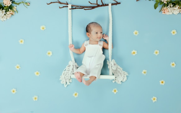 A baby dressed in a white outfit lies on a decorative swing made of white ropes and a wooden branch. The background is light blue with scattered small white flowers. Green leaves and flowers decorate the top corners, enhancing the whimsical and serene atmosphere.