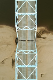 An overhead view of a bridge with a geometric lattice structure spanning over a body of water with sandy shores. The bridge has a light blue metallic frame and a concrete roadway.
