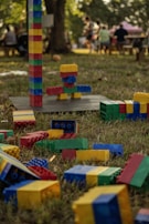 Colorful building blocks are scattered on the grass with a tall tower constructed from similar blocks nearby. In the background, people are gathered casually under a tree, possibly enjoying an outdoor event.