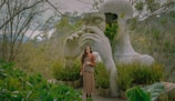 A large, artistic sculpture of a human head integrated with lush greenery in a peaceful, natural setting. A person stands in front of the sculpture, surrounded by various plants. The scene conveys a harmonious blend of nature and art.