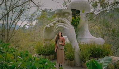 A large, artistic sculpture of a human head integrated with lush greenery in a peaceful, natural setting. A person stands in front of the sculpture, surrounded by various plants. The scene conveys a harmonious blend of nature and art.