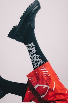 A leg is raised, showcasing a black chunky shoe paired with a black sock featuring white lettering. The wearer is dressed in red pants with white abstract patterns against a neutral background.