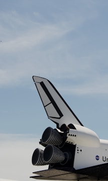 The lower rear section of a space shuttle is visible, featuring three large rocket engines and a vertical tail fin. The shuttle exterior is white with black details, and a NASA logo is partially visible. A clear blue sky forms the background.