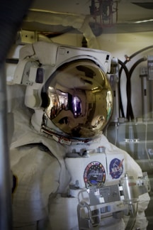 An astronaut wearing a white space suit with various patches including a NASA logo, inside a spacecraft or space station. The helmet visor is reflective and shows a distorted reflection of the interior environment.