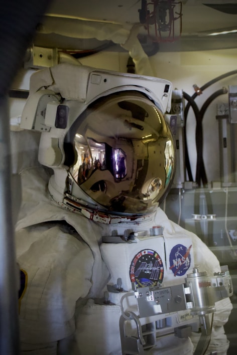 An astronaut wearing a white space suit with various patches including a NASA logo, inside a spacecraft or space station. The helmet visor is reflective and shows a distorted reflection of the interior environment.
