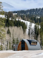 A sleek, steel-structured prefabricated cottage nestled on a snowy slope with burnt orange accents.