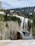 A modern, arched cabin with a sleek design is situated in a snowy landscape, surrounded by dense coniferous and bare deciduous trees. Snow covers the ground and the cabin is backdropped by a forested hillside under a partly cloudy sky.