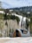 A modern, arched cabin with a sleek design is situated in a snowy landscape, surrounded by dense coniferous and bare deciduous trees. Snow covers the ground and the cabin is backdropped by a forested hillside under a partly cloudy sky.