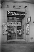 A storefront of a deli or food shop with the name 'Don Jamón' displayed above the entrance. The glass door shows reflections and various posters or images featuring food items. A small menu or sign is placed near the entrance, and a chair is positioned beside the door.
