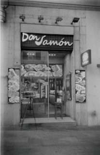 A storefront of a deli or food shop with the name 'Don Jamón' displayed above the entrance. The glass door shows reflections and various posters or images featuring food items. A small menu or sign is placed near the entrance, and a chair is positioned beside the door.