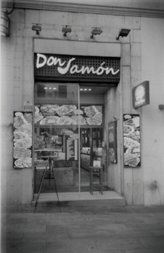 A storefront of a deli or food shop with the name 'Don Jamón' displayed above the entrance. The glass door shows reflections and various posters or images featuring food items. A small menu or sign is placed near the entrance, and a chair is positioned beside the door.