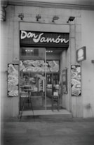 A storefront of a deli or food shop with the name 'Don Jam&oacute;n' displayed above the entrance. The glass door shows reflections and various posters or images featuring food items. A small menu or sign is placed near the entrance, and a chair is positioned beside the door.