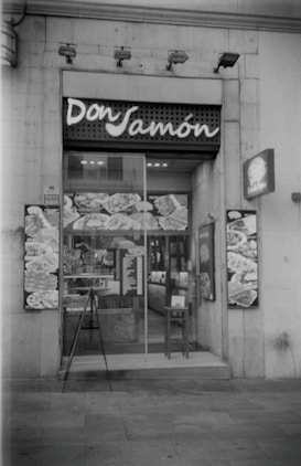 A storefront of a deli or food shop with the name 'Don Jamón' displayed above the entrance. The glass door shows reflections and various posters or images featuring food items. A small menu or sign is placed near the entrance, and a chair is positioned beside the door.