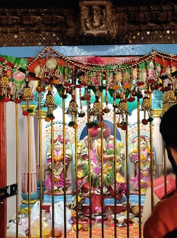 The image depicts a colorful shrine with intricate decorations. It features a statue of a deity adorned with flowers and decorations. In front of the shrine, there are offerings placed on the ground including fruits and bags, surrounded by a decorative barrier with bells and ornaments, reflecting vibrant festival colors.