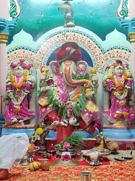A vibrant statue of the Hindu deity Ganesha is adorned with colorful garlands, surrounded by two smaller statues on either side. The figures are richly decorated with traditional garments and jewelry, set against an intricately painted backdrop. Offerings, flowers, and ceremonial objects are placed in front of the statues on a red cloth-covered altar.