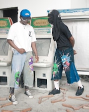 Two individuals stand in front of vintage arcade machines. They are wearing uniquely painted denim pants with expressive, colorful designs, such as vibrant cartoon-like faces. One person wears a white t-shirt and blue cap, while the other has a black shirt with a head covering. The floor is scattered with wooden crutches, creating a chaotic atmosphere.