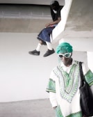 Two individuals are in a minimalistic, unfinished concrete setting. One person is sitting on an elevated platform, partially obscured, wearing dark clothing, with their face covered. The other person stands below, dressed in a vibrant, patterned green and white outfit with headwrap and sunglasses, holding a black bag.