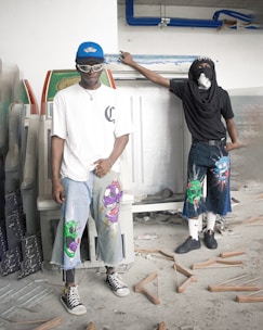 Two individuals are standing in what appears to be a cluttered space, possibly a workshop or storage area. Both are dressed in unique, urban streetwear featuring cartoonish designs on their shorts. One is wearing a blue cap and sunglasses, while the other has a mask covering their face. The floor is scattered with wooden coat hangers, adding to the disorganized setting.