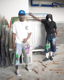 Two individuals are standing in what appears to be a cluttered space, possibly a workshop or storage area. Both are dressed in unique, urban streetwear featuring cartoonish designs on their shorts. One is wearing a blue cap and sunglasses, while the other has a mask covering their face. The floor is scattered with wooden coat hangers, adding to the disorganized setting.