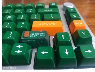 A close-up view of a keyboard with mostly green keys, featuring distinctive orange keys labeled 'ACTIVATE' and 'STOP'. The green keys are arranged with various symbols, and there is a component labeled 'PB 08C300 CA07 006' visible among the keys.