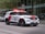 A police SUV is driving along a city street. The vehicle is white with red accents and equipped with visible emergency lights on the roof and front grille. The background features modern glass buildings and a couple of parked vehicles.