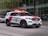 A police SUV is driving along a city street. The vehicle is white with red accents and equipped with visible emergency lights on the roof and front grille. The background features modern glass buildings and a couple of parked vehicles.
