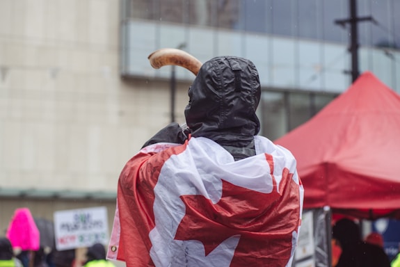 A person wearing a black hooded jacket is draped in a Canadian flag while holding a horn-like object. The background includes a red canopy tent and indistinct signs with some text visible. It appears to be a rainy day, with droplets visible in the air.
