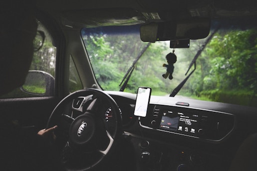 The interior of a car is visible with a person driving. The dashboard features a GPS device mounted on it, and the windshield wipers are in motion as it rains outside. The rearview mirror has a small fuzzy ornament hanging from it, and greenery is visible through the windows.