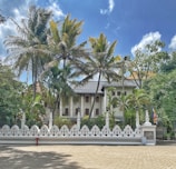 A colonial-style building is partially obscured by tall palm trees, surrounded by a decorative white fence with geometric patterns. The sky is bright with scattered clouds, providing a serene backdrop. To the side, a flag in vibrant colors is visible.