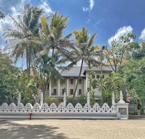 A colonial-style building is partially obscured by tall palm trees, surrounded by a decorative white fence with geometric patterns. The sky is bright with scattered clouds, providing a serene backdrop. To the side, a flag in vibrant colors is visible.