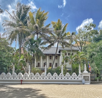 A colonial-style building is partially obscured by tall palm trees, surrounded by a decorative white fence with geometric patterns. The sky is bright with scattered clouds, providing a serene backdrop. To the side, a flag in vibrant colors is visible.