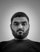 A black and white portrait of a man with a beard and short hair. He is seen from a low angle and wears a serious or contemplative expression. The background is plain and free of distractions.