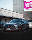 A sleek black Honda Civic Type R is parked in a commercial area. The car's front end features a prominent red Honda emblem and sporty design elements. In the background, there is a large sign with the words 'South Flatz' in bold pink and white letters against an industrial building.