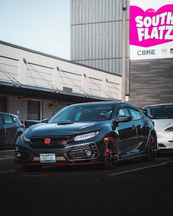 A sleek black Honda Civic Type R is parked in a commercial area. The car's front end features a prominent red Honda emblem and sporty design elements. In the background, there is a large sign with the words 'South Flatz' in bold pink and white letters against an industrial building.