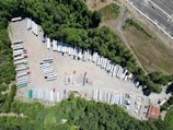 Aerial view of a truck yard with numerous parked trailers and vehicles, surrounded by dense greenery. The area includes various colored trailers and a few small buildings along the edge.