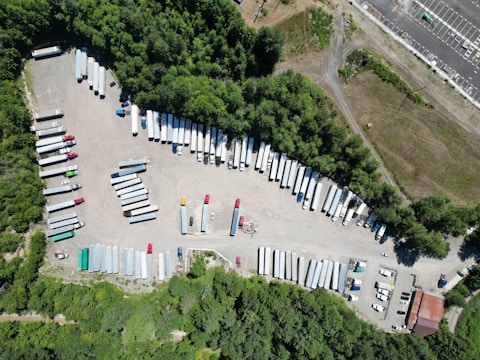 Aerial view of a truck yard with numerous parked trailers and vehicles, surrounded by dense greenery. The area includes various colored trailers and a few small buildings along the edge.