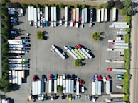 An aerial view of a parking lot filled with trucks and trailers arranged in rows. The vehicles are organized in a systematic manner, with some smaller cars and trailers mixed in. Trees and small green spaces are distributed throughout the area, providing some greenery amid the concrete.